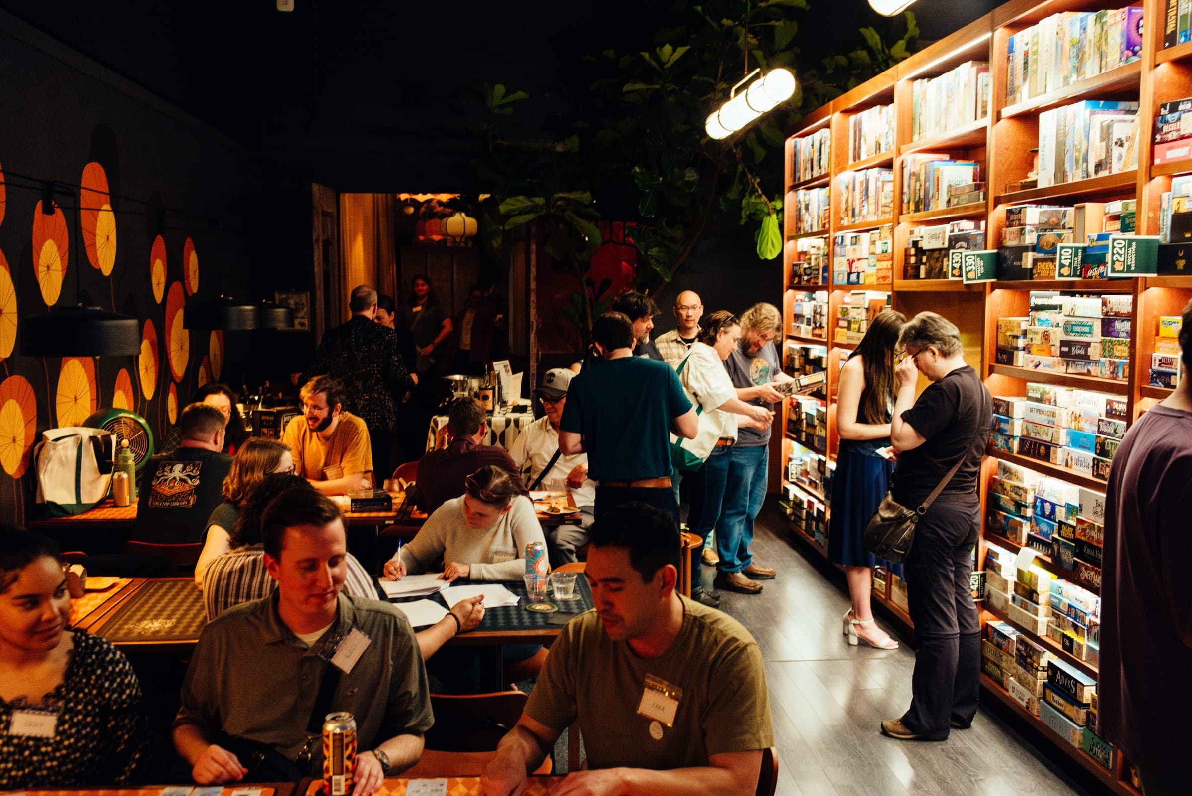 A full room on an event night, with players at the tables and people browsing the lit shelves of games